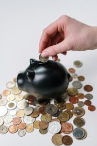 pexels-photo-3943723-3943723 Hand inserting coin into a black piggy bank surrounded by various coins, symbolizing savings.